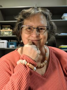 Older woman with shades of coral in jewelry and sweater, glasses, silver and brown hair.