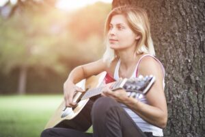 young woman with blonde hair sitting at a tree playing guitar