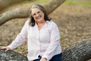 60 year old woman, with dark hair and a white streak in front, in a pink blouse, sitting on a tree branch, looking at the camera